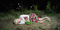 Eine Frau liegt in einem Rosenkleid auf einem Feld, sie greift zu einer Gurke, dahinter Blumen