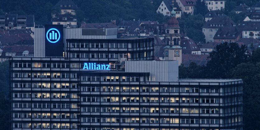 Großes, kastiges Gebäude mit blau leuchtendem Logo der Allianz auf dem Dach bei Nacht.