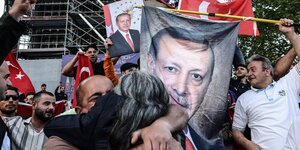Menschen umarmen sich und schwingen türkische Fahne vor einem Plakat des türkischen Präsidenten