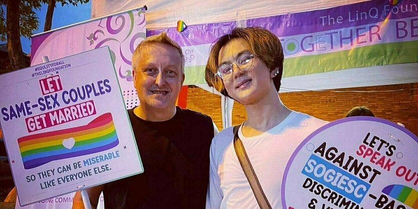 Zwei Männer stehen vor einer Regenbogenfahne und halten Schilder in den Händen