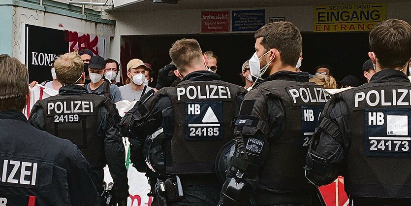 Das Bild zeigt fünf Polizist*innen von hinten, die einer Reihe von Demonstrierenden gegenüberstehen