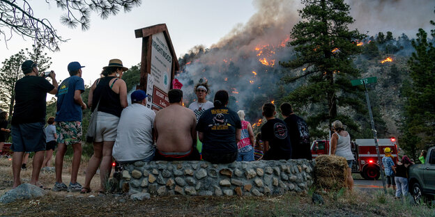 Evakuierte Menschen sitzen auf einer Mauer. Dahinter ist ein großer Waldbrand zu sehen