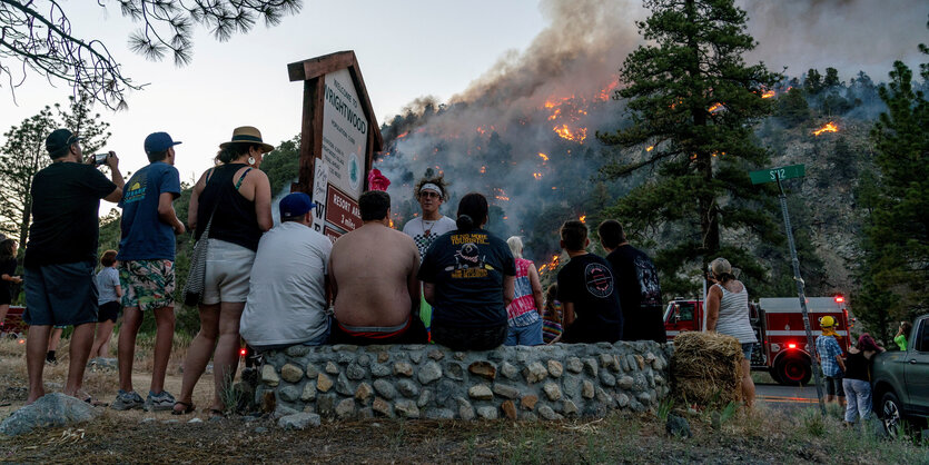 Evakuierte Menschen sitzen auf einer Mauer. Dahinter ist ein großer Waldbrand zu sehen