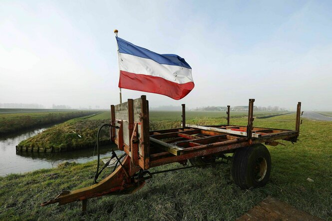 Auf einem Traktoranhänger, der auf einer Wiese vor einem Kanal steht weht die auf den Kopf gestellte niederländische Flagge