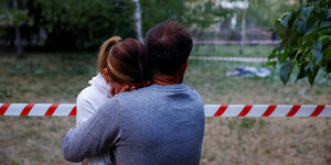 Ein Mann umarmt eine Frau. Sie stehen vor einer Absperrung und schauen auf einen toten Körper (der aber nicht sichtbar auf dem Bild ist).