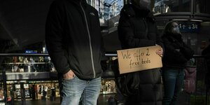 Zwei junge Menschen stehen auf dem Bahnhof, dei Frau trägt ein Schild: We offer 2 free beds