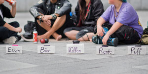 Punks sitzen auf der Straße, vor sich mehrere Becher für Kleingeld mit den Aufschriften "Essen", "Bier", "Kiffen", "Rente" und "Puff".
