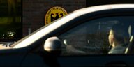 Eine Frau sitzt in einem Auto, hinter ihr sieht man das Schild einer deutschen Botschaft