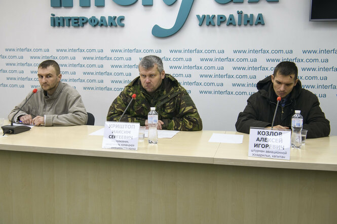 Pressekonferenztisch in Kyiw, dahinter sitzen drei Männer, einer ist der gefangengenommene russische Kampfpilot Krishtop