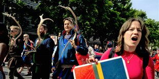 Menschen, teilweise in samischer Tracht gehen auf einer Allee entlag. Die erste Reihe ist aus nächster Nähe fotografiert. Drei Menschen links halten Rentiergeweihe in den Händen. Eine vierte Person mit rotem Oberteil und einem Plakat in den Farben der samischen Flagge hat den Mund weit geöffnet, als würde sie etwas rufen.