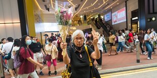 Eine Frau mit weißem Haar hält einen Strauß mit roten Lilien in die Höhe und macht mit der anderen Hand ein Zeichen