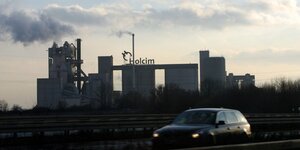 Ein Auto fährt auf einer Autobahn vor einer Fabrik der Firma Holcim, die Schornsteinschlote rauchen