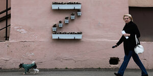 Eine Frau führt ihren Hnd spazieren, sie geht an einer Hauswand mit Blumentöpfen vorbei, die zu einem "Z" arrangiert sind