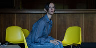 Die Musikerin Caterina Barbieri sitzt vor einer Holztheke seitlich nach rechts zur Kamera gewandt auf einem gelben Schalenstuhl, neben ihr stehen zwei weitere gelbe Stühle. Sie trägt eine Hose und ein Hemd aus blauem Jeansstoff und blickt in die Kamera.