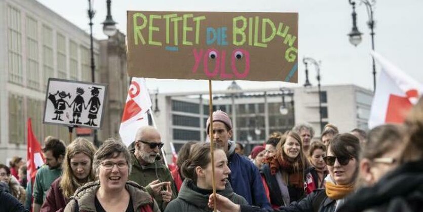 Die Gesichter von vielen menschen sind zu sehen, die für die GEW streiken. Über ihren Köpfen wird ein Schild gehalten. Die Aufschrift: "Rettet die Bildung, yolo".