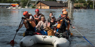 Sieben Menschen und ein Hund sitzen in einem Schlauchboot. Vier von ihnen paddeln mit Rudern. Im Hintergrund sieht man überschwemmte Häuser