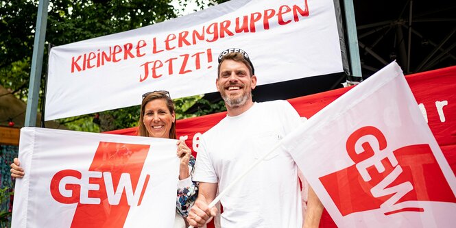 Zwei Lehrer, ein Mann und eine Frau, mit GEW-Fahnen und vor dem Transparent "Kleinere Lerngruppen jetzt"