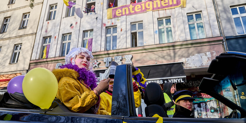 Eine Frau in den Farben gelb und lila der Kampagne sitzt in einem geschmückten offenen Auto, das an einem Haus mit "Enteignen"-Banner vorbeifährt