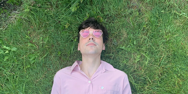 Ein Mann in rosa Hemd, braunen Haaren und einer Brille mit Brillengläsern liegt mit geschlossenen Augen, deren Lider durch die rosa-herzförmigen Brillengläser zu sehen sind, auf grünem Rasen.