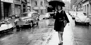 Eine lächelnde Frau geht mit einem Regenschirm über dem Kopf eine regnerische Straße entlang