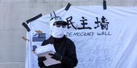 Ein vermummter Mensch steht vor einer weißen Wand, auf der "Democracy Wall" steht und chinesische Schriftzeichen