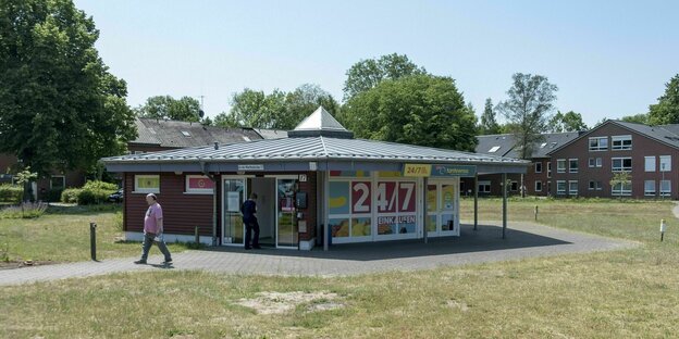 Ein TanteEnso Laden - typischer kleiner Zweckbau - steht auf einem Dorfanger - auf der Fassade steht 24/7 -Kunde betritt den Laden, einer geht weg