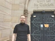 Person mit schwarzem Pulli und Brille vor einer schwarzen Kirchentür