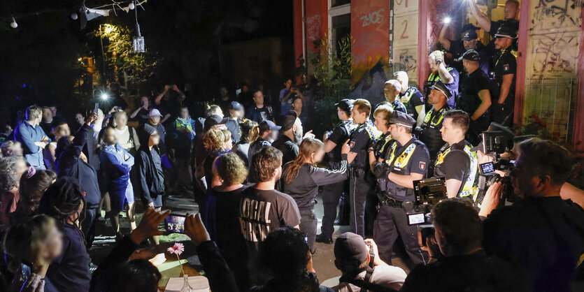 Aufgebrachte Menschen und Polizei nachts vor einer Tür, die Polizei steht im Türeingang