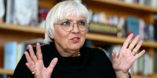 Porträt Claudia Roth, dunkler Pulli, Brille, lächelt und gestikuliert mit Händen vor Regalwand