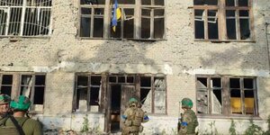 Soldaten stehen vor einem zerstörtem Gebäude. Aus einem Fenster weht die ukrainische Flagge
