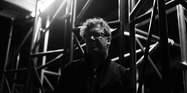 Schwarz-weißes Porträtfoto von Hanno Leichtmann. Der Musiker trägt ein Hemd und eine viereckige Brille. Er steht vor einem Gerüst, das geometrische Schatten wirft.