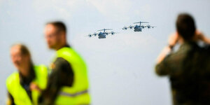 Zwei Flugzeuge der Luftwaffe am Himmel, im Vordergrund sind unscharf Menschen zu erkennen. Zwei Männer tragen gelbe Warnwesten, ein dritter Mann schaut durch ein fernglas