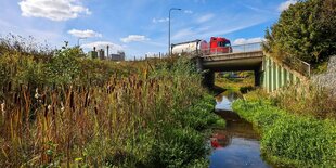 Ein LKW fährt über eine Brücke die über einen renaturierten Bach führt.