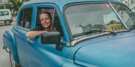 Eine junge Frau lehnt sich lächelnd aus dem geöffneten Beifahrerfenster eines blauen Taxis