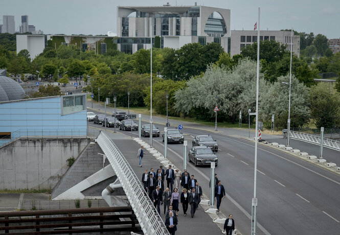 Bundeskanzler und Bundesminster:innen gehen auf einer Straße. Hinter ihnen fährt eine Sicherheitseskorte schwarzer Autos.