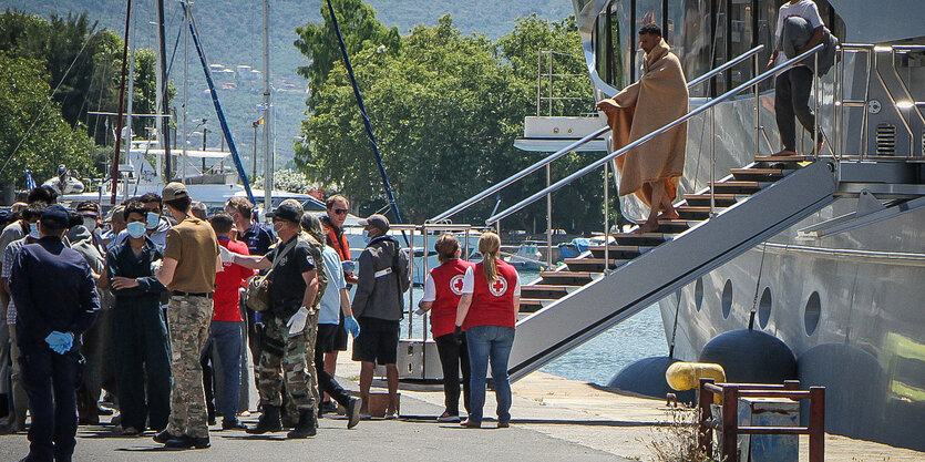 Menschen stehen in Decken gehüllt auf einem Kai, dahinter die Stiege zu einem Schiff