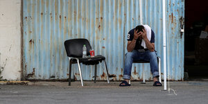 Ein Mann stützt seinen Kopf auf die Hände, er sitzt auf einem Stuhl vor einer Lagerhalle aus Wellblech