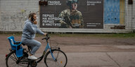 Frau fährt Fahrrad und telefoniert, im Hintergrund Kriegsplakat in der Ukraine