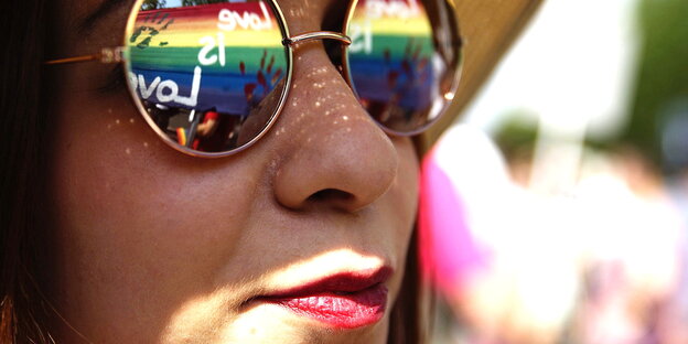 Frau mit Hut und Sonnenbrille, auf deren Gläsern sich die Regenbogenflagge spiegelt