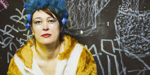 Patrica Wedler mit blauer Perücke und in einem Plüschanzug vor einer mit Graffiti bemalten Tafel