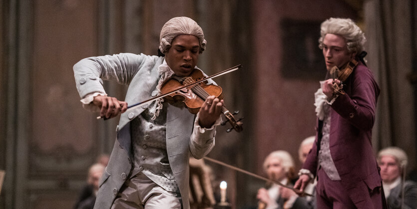 Das Standphoto zeigt links Joseph Bologne, der gerade Violine spielt. Rechts hört Mozart zu und ahnt wohl schon, dass er diesen Wettbewerb verlieren wird