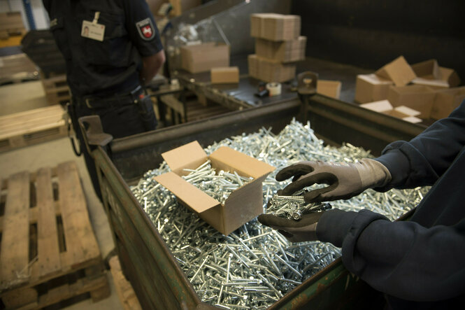 Schrauben werden von einer Hand mit Schutzhandschuhen auf einer großen Kiste in eine Pappschachtel gepackt