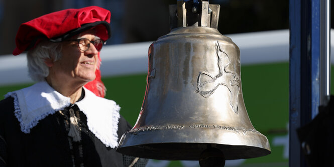 Ein Mann in einem historischen Kostüm steht neben einer Glocke mit Friedenstaube.