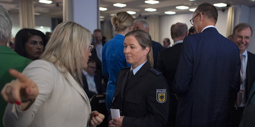 Pechstein in Polizeiuniform spricht mit einer Frau. An ihnen geht Merz vorbei