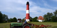 Im Vordergrund ein Traktor auf einer grünen Wiese, im Hintergrund ein weiß-rot geringelter Leuchtturm.