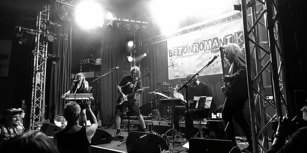 Schwarz-weiße Aufnahme von links nach rechts: Die Band Rosa Beton auf der Bühne: Martina Dünkelmann steht am Keyboard und singt, Thomas Wagner spielt E-Gitarre, Alexander Lode sitz an einem Drumcomputer, hinter ihm steht ein Schlagzeug, und Thomas Trenkel spielt Bass.