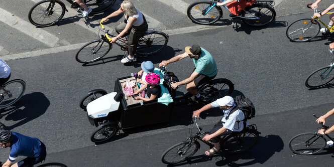 Eine Lastenrad mit 2 Kindern beladen und viele Räder drumherum brausen auf einer Straße