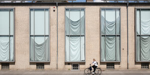 Blick von außen auf das Gebäude der Arthur-Boskamp-Stiftung: fünf hohe rechteckige Fenster, mit weißem Stoff verhängt; davor fährt eine Fahrradfahrerin vorbei