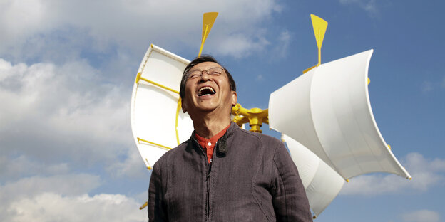 Der Künstler Susumu Shingu steht vor einer kinetischen Skulptur aus gelben Stahlträgern und weißen Stoffsegeln. Dahinter ist der blaue Himmel mit Wolken zu sehen. Der Künstler trägt eine graue Jacke, darunter ein orangenes Hemd und eine Brille. Er hebt de