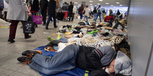 ein Mann liegt auf einer Decke auf dem Boden in einer Bahnhofshalle, an ihm vorbei laufen Passanten